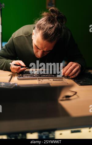 Vertical portrait of focused technician examining and repairing laptop motherboard, troubleshooting hardware issues sitting at table in workshop. Stock Photo