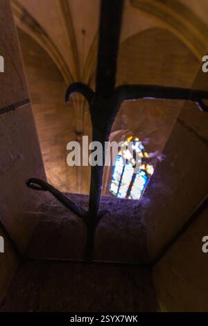 A low-angle shot looking up into a stone spiral staircase with a stained glass window at the top, framed by the curving steps and ornate ironwork. Stock Photo