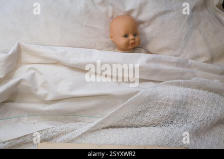 A doll lies in a bed, likely a historical hospital bed, draped in white linens.  Possible display at a museum. Stock Photo