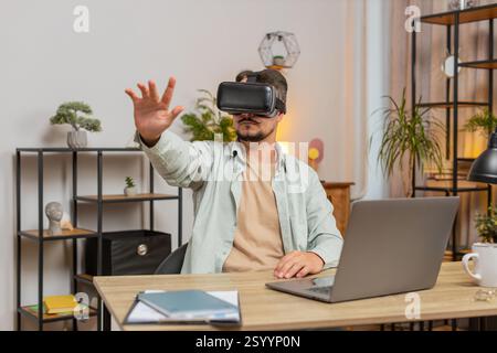 Confident Caucasian man freelancer testing VR headset while sitting at home office table. Young businessman guy making presentation using new gadget during remote working online at home. Lifestyle. Stock Photo