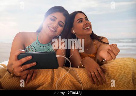 Two female friends listening to music while lying down on beach Stock Photo