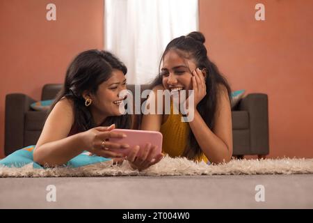 Two smiling women together watching video on mobile phone while lying on floor in living room Stock Photo