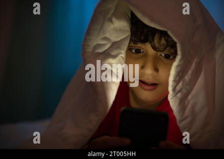 Boy using mobile phone under bed cover at night Stock Photo