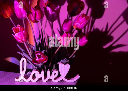 Wooden word LOVE on background of Pink colored tulip flower in neon light on violet gradient background in the night light. Vale Stock Photo