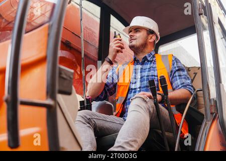 Young man loader worker or a forklift driver in a container warehouse wearing a white helmet is talking on the walkie-talkie or radio phone. Stock Photo