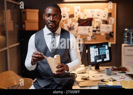 Waist up portrait of Black young man as detective eating takeout noodles in office and looking at camera, copy space Stock Photo