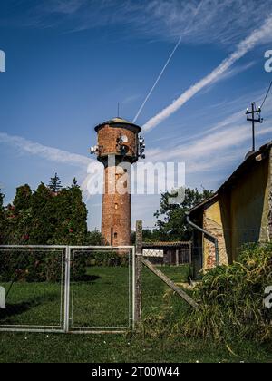 Brick old water tower with telecommunications equipment in a rural setting next to a farm green field tree blue cloudy sky Stock Photo