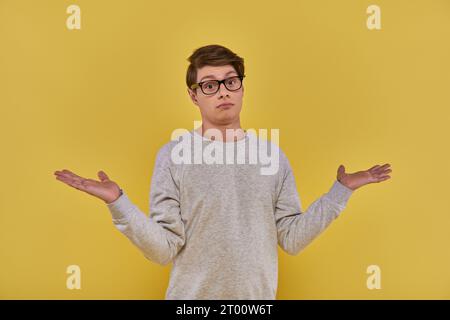 confused clueless young man in sweatshirt and glasses showing helpless gesture on yellow backdrop Stock Photo