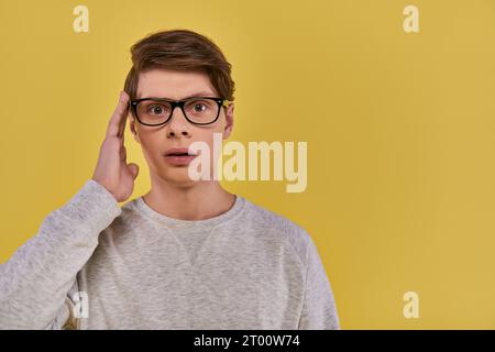 shocked man in white sweatshirt touching glasses and slightly opening his mouth on yellow backdrop Stock Photo