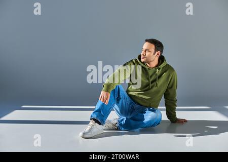 indian man in casual street wear sitting on floor and looking away on grey background Stock Photo