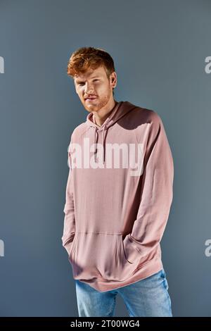 stylish young red haired man in pink hoodie and jeans looking at camera posing on grey backdrop Stock Photo