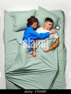 Young beautiful woman reaching across attractive man lying on back for turning of loud alarm clock, couple relaxing in big mint bed. Overslept. Stock Photo