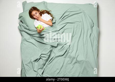 Top view portrait of little girl lying on back covered with blanket surprised looking at phone. Lovely kid, child in big mint bed. Stock Photo