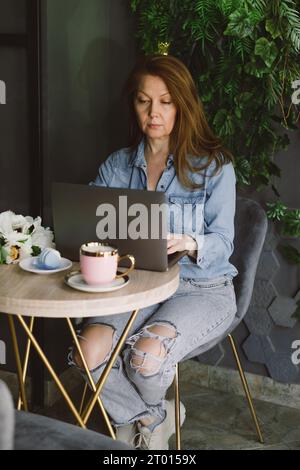 Woman working on laptop in cafe. Middle-aged freelancer with cup of coffee typing and reading. Loft working place with green plants Stock Photo