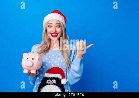 Photo portrait of lovely blonde young lady hold piggy point empty space wear penguin sweater x-mas hat isolated on blue color background Stock Photo