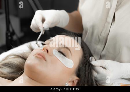 A beautician looks in a mini mirror and looks at the newly extended eyelashes of a young woman in a beauty salon. Woman's face in cosmetic procedure Stock Photo
