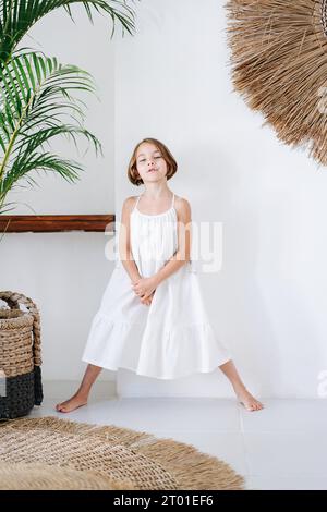 A child in a white dress, legs spread wide, against the background of a white wall of a summer house Stock Photo