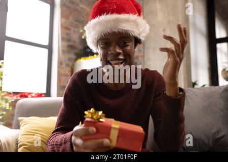 Happy african american man at home in christmas hat having video call, holding gift and talking Stock Photo