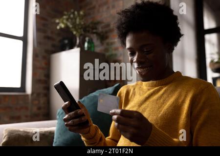 Happy african american man at home holding credit card and using smartphone to make online payment Stock Photo