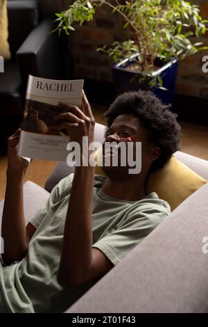 Laughing african american man lying on couch reading book wearing under eye masks Stock Photo