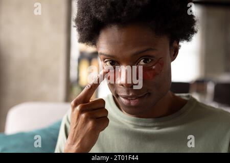 Portrait of african american man sitting on couch at home applying under eye masks Stock Photo