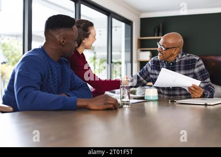 Diverse couple and biracial male financial advisor using laptop and shaking hands at home Stock Photo