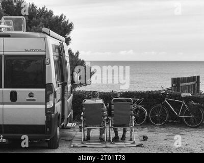 Scenic RV Campsite Pitch, Camper Van in the recreational vehicles park, Woman and man relaxing on a chair and watching Sunset Over the sea Stock Photo