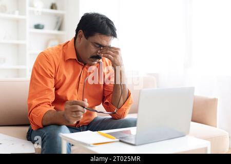 Stressed mature businessman sitting at workplace, holding eyeglasses, rubbing eyes Stock Photo