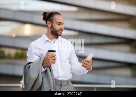 Handsome young businessman using smartphone and drinking coffee while walking outdoors Stock Photo