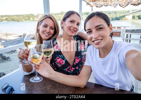 Three cheerful girlfriends sit at a table with a glass of wine drinking, talking, and taking selfies on a restaurant terrace by the river. Stock Photo