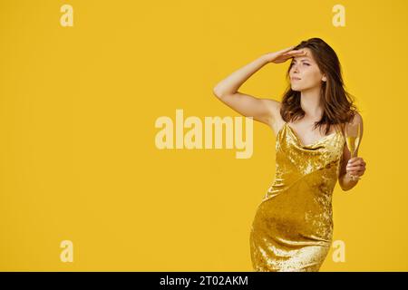 Beautiful woman in evening dress holding glass of champagne and looking aside at empty space, yellow studio background Stock Photo