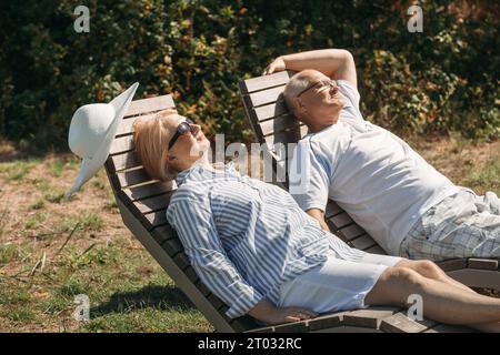 A couple of cute elderly people lie on sunbeds holding hands, basking in the sun. Stock Photo