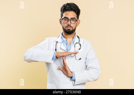 Tired serious upset Indian young doctor cardiologist man showing time out gesture, limit or stop sign, take a break, relax, rest, help. Arabian apothecary pharmacy guy isolated on beige background Stock Photo
