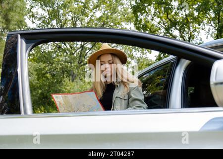 Blonde woman in hat staying next to car door checks the route on map to get to the destination. Young tourist explore local travel making candid real moments. True emotions expressions of getting away and refresh relax on open clean air Stock Photo