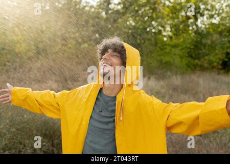 Smiling arabic man wearing a yellow raincoat during the rain in the park. Handsome male enjoying the rain outdoors. Guy has a joyful expression during Stock Photo
