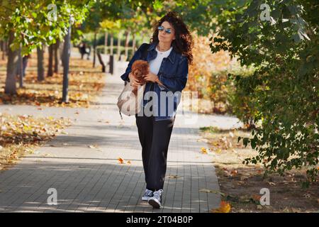 Full length stylish curly brunette woman walks in park and holds dog in bag. Stroll with pet in city. Stock Photo