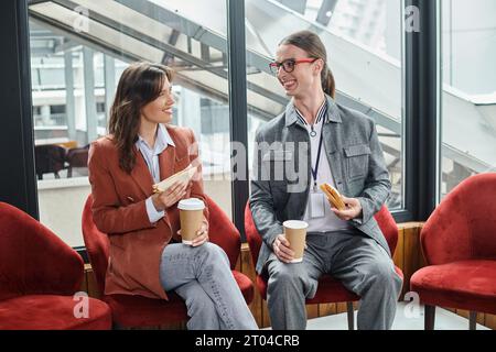 two coworkers in smart casual attire sitting in chairs chatting and eating, coworking concept Stock Photo