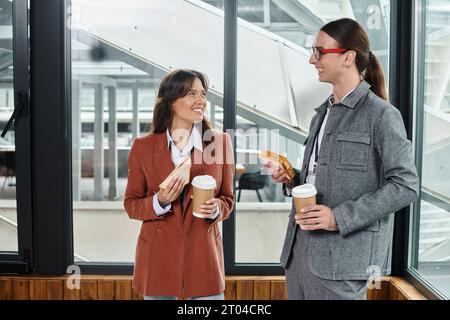 two young colleagues having sandwich with tea on break and smiling at each other, coworking concept Stock Photo