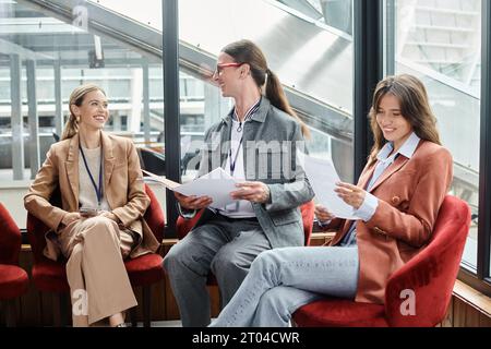 three cheerful team members sitting on red chairs and discussing paperwork, coworking concept Stock Photo