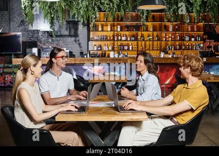 hardworking cheerful team using laptops and smiling to each other with blurred backdrop, coworking Stock Photo