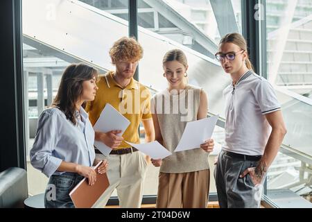 four young colleagues focused on examining working papers with window on backdrop, coworking Stock Photo