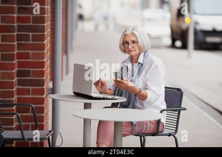 Gray haired elderly businesswoman sitting in cafe on street with mobile phone and laptop, senior woman working using technology and gadgets. Stock Photo