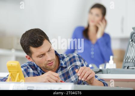 serviceman testing appliance with multimeter housewife in background Stock Photo