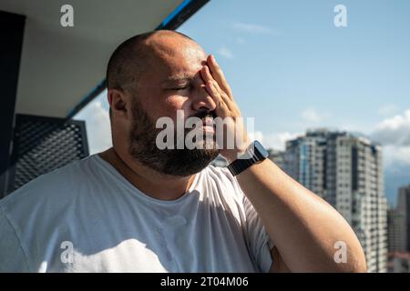 Fat man suffers dying from heat. Guy on balcony under hot summer sun sweats from high temperatures Stock Photo