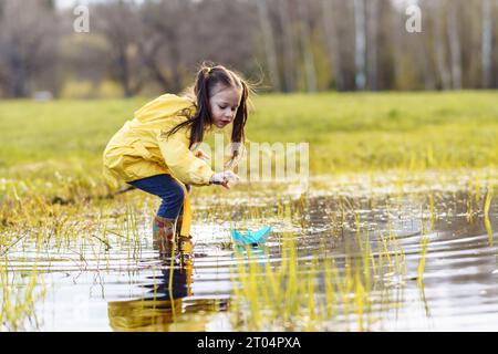 Charming little girl in yellow jacket and rubber boots standing in puddle on green lawn, stretching arm forward and bending over, carefully looking at Stock Photo