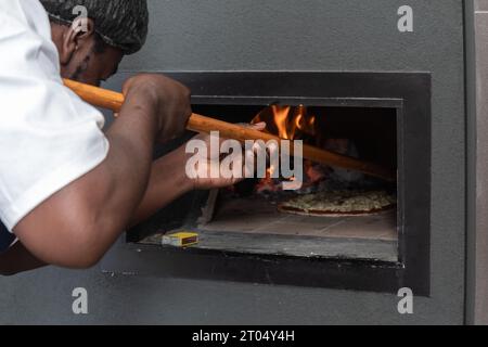 In the intense glow of the pizza oven's flames, a skilled black man, using a wooden peel, expertly maneuvers a sizzling pizza Stock Photo