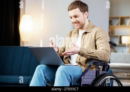 Handsome man with a disability sitting in wheelchair with laptop on knees and looking at camera. Caucasian male in casual wear having opportunities to Stock Photo