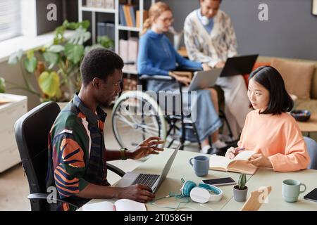 Colorful portrait of two young professionals talking while collaborating on project at meeting table Stock Photo