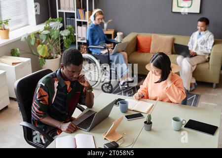 High angle portrait of Black young man using laptop computer in office with diverse team of young professionals, copy space Stock Photo