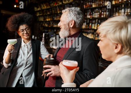 bearded middle aged man smiling near multiethnic women with cocktails, colleagues resting in bar Stock Photo